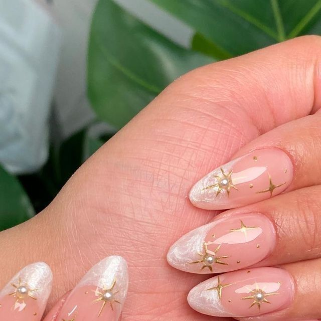 Chic Almond-Shaped Nails: Nude Base, Delicate White Tips, and Glamorous Gold Starbursts with Pearls.