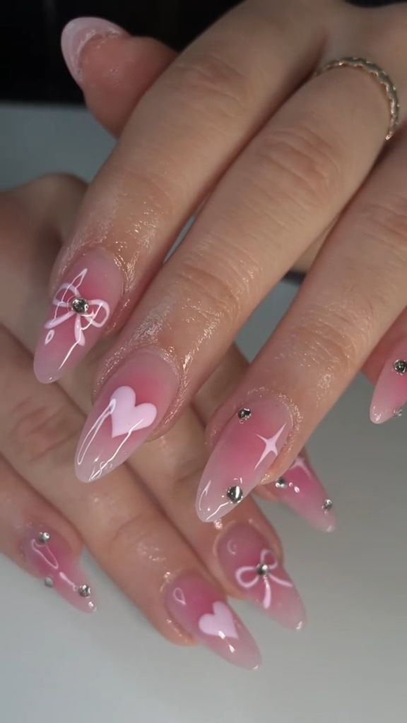 Charming Soft Pink Gradient Nail Art with White Patterns, Heart and Bow Motifs, Enhanced by Sparkling Rhinestones.