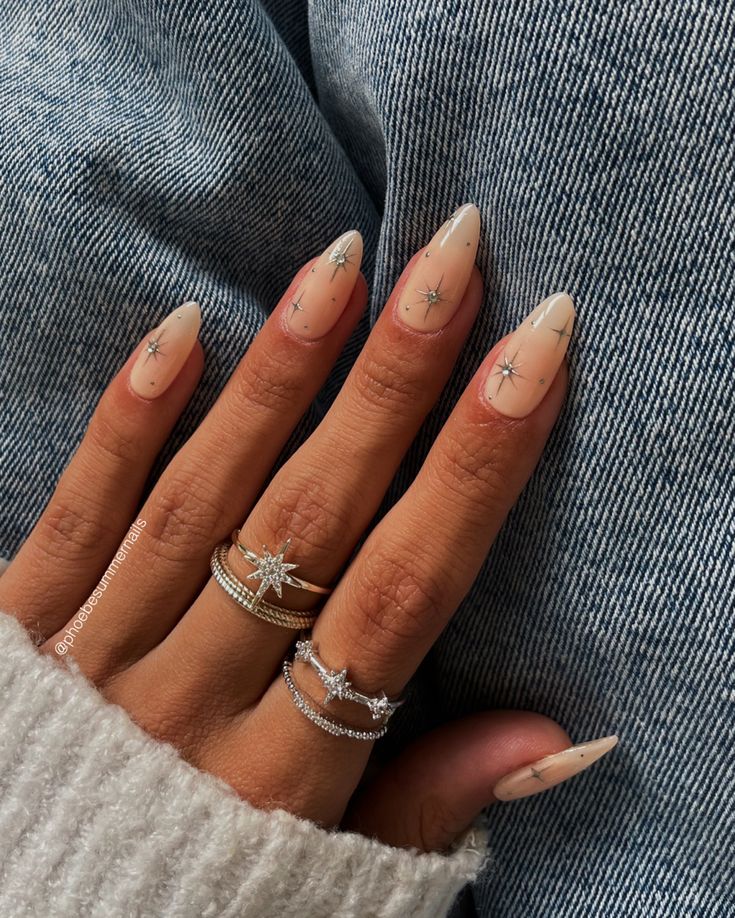 Chic Almond-Shaped Nails with Soft Nude Base and Delicate Star Motifs, Enhanced by Intricate Silver Jewelry.