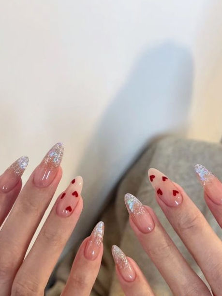 Chic Almond-Shaped Nail Design: Nude Base with Red Hearts and Glitter Tips for a Glamorous Look.