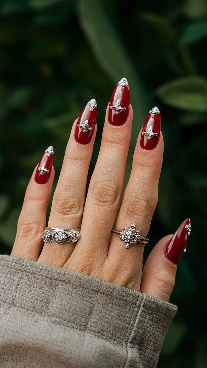 Elegant Red Manicure: Glossy Almond-Shaped Nails Enhanced with Intricate Silver Embellishments for Glamour and Sophistication.