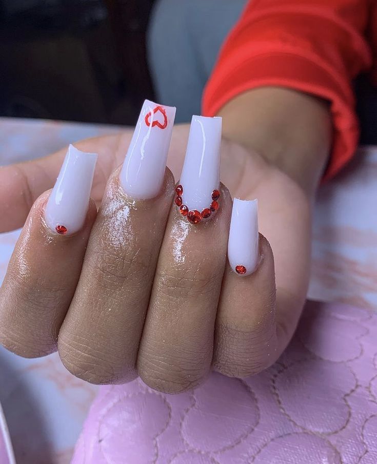 Chic Glossy White Acrylic Nails with Intricate Red Accents and Sparkling Rhinestones.