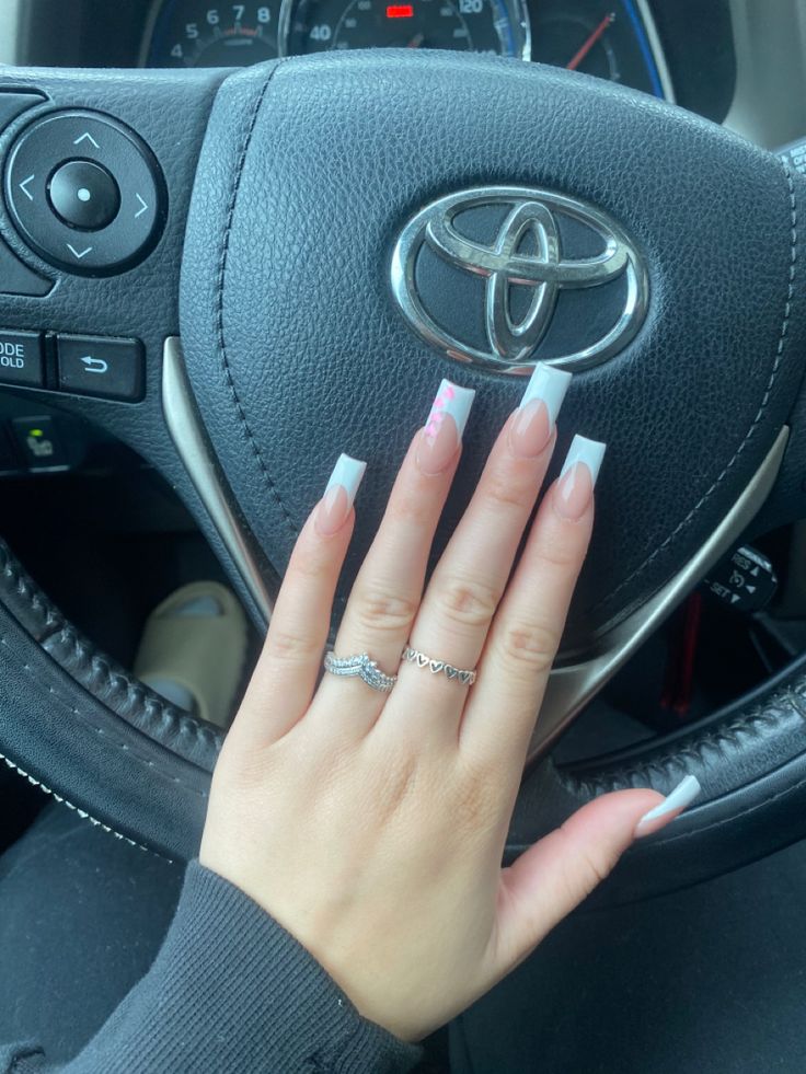 Chic Long Acrylic Nails with French Tips and Color Accent, Enhanced by Sparkling Rings and Car Background.