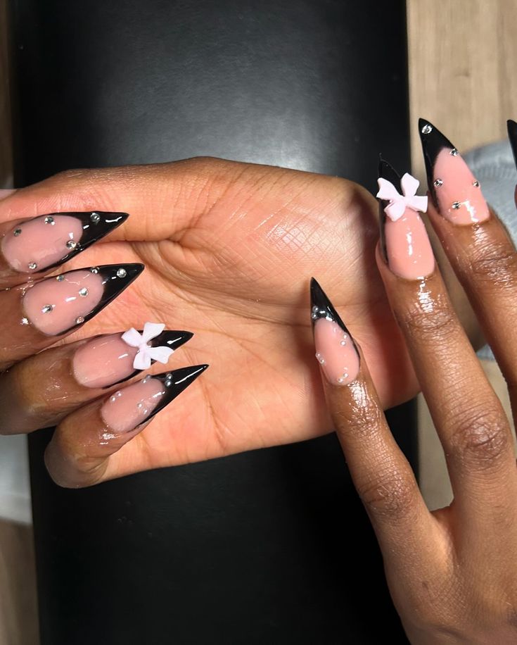 Chic Elongated Stiletto Nail Design with Black Tips, Pastel Pink Accents, Rhinestones, and White Floral Embellishments.