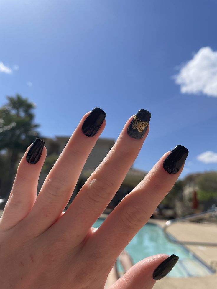 Chic Black Nails with Elegant Gold Butterfly Accent in a Vibrant Outdoor Setting.