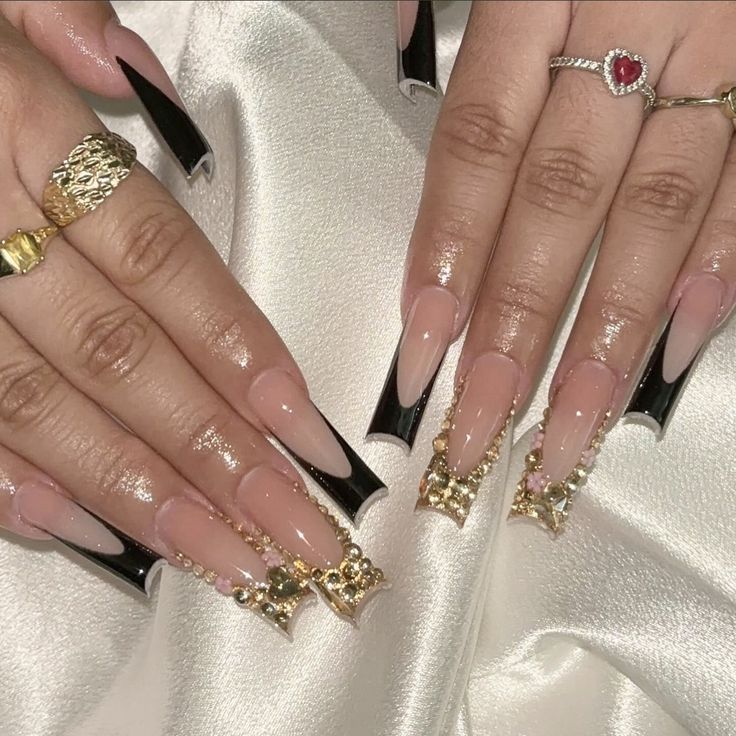 Chic Stiletto Nail Design with Nude Base, Bold Black Accents, and Luxurious Gold & Jewel Embellishments.