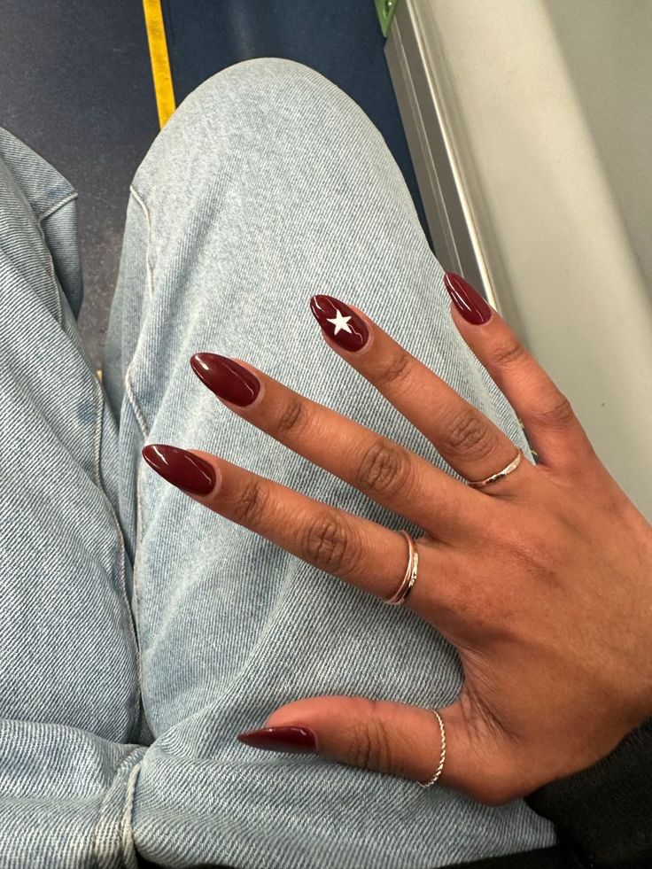 Sophisticated Burgundy Almond Nails with Striking White Star Design and Minimalist Silver Rings.