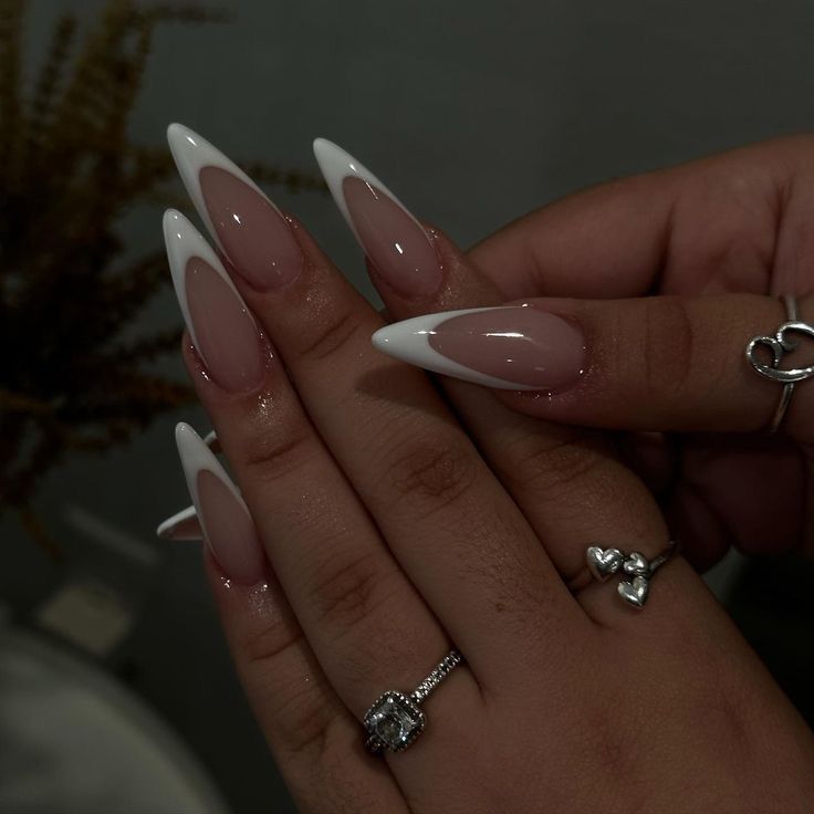 Elegant Stiletto Nails: Chic Pale Nude and Bold White Tips with Glossy Finish and Silver Rings.