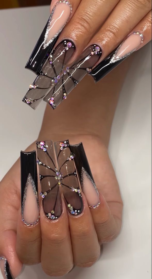 Chic Manicure Design: Long Sharp Nails in Black and Nude with Floral Patterns and Gemstone Embellishments.