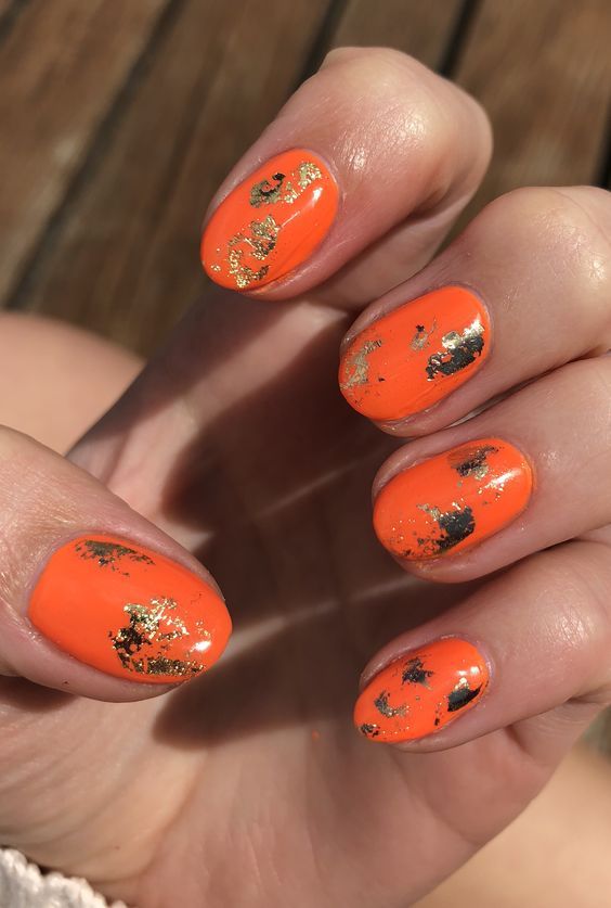 Striking Vibrant Orange Nail Design with Gold Flakes: A Perfect Blend of Elegance and Playfulness for Any Occasion.