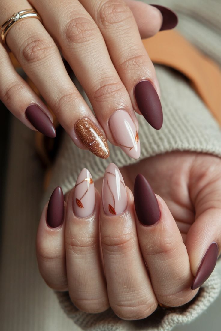 Sophisticated Nail Design Featuring Matte Burgundy, Sparkly Gold Accents, and Delicate Leaf Patterns on Nude Bases.