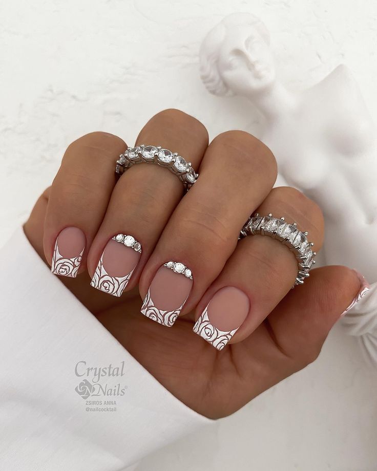 Chic Nude Nail Design with Floral Tips and Sparkling Rhinestones Accented by Silver Rings.