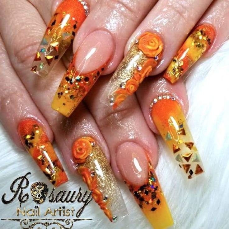 Glamorous Orange and Gold Stiletto Nail Design with Floral Accents and Rhinestones.