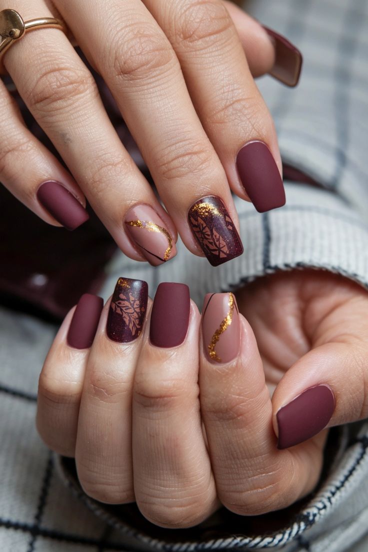 Sophisticated Burgundy and Nude Nail Design with Leaf Patterns and Gold Accents.