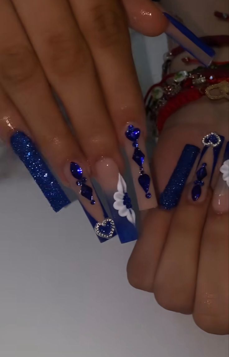 Elegant Striking Blue Nail Design with Glitter, Floral Accents, and Gem Embellishments.