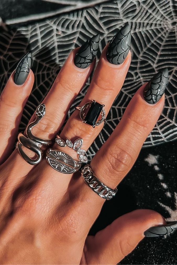 Bold Black Stiletto Nails with Spiderweb Designs and Silver Rings Create a Chic, Edgy Statement Look.