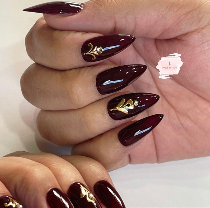 Sophisticated Maroon Almond Nails Embellished with Gold Accents and Gems for Glamorous Occasions.