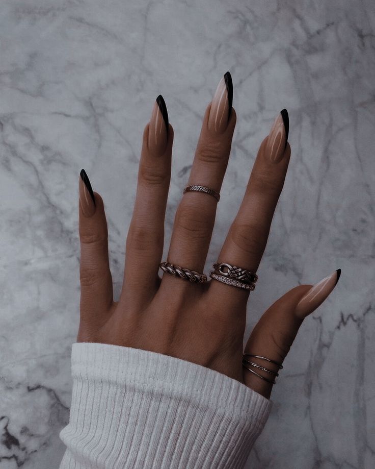 Chic Almond-Shaped Nails with Nude Base and Black Tips Complemented by Delicate Rings in a Minimalist Setting.