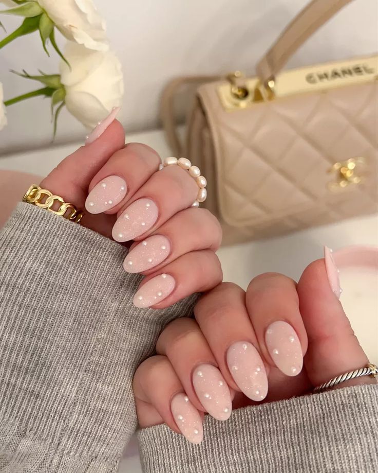 Chic Almond-Shaped Nails: Soft Nude with Playful White Polka Dots, Styled with Grey Sweater and Elegant Handbag.