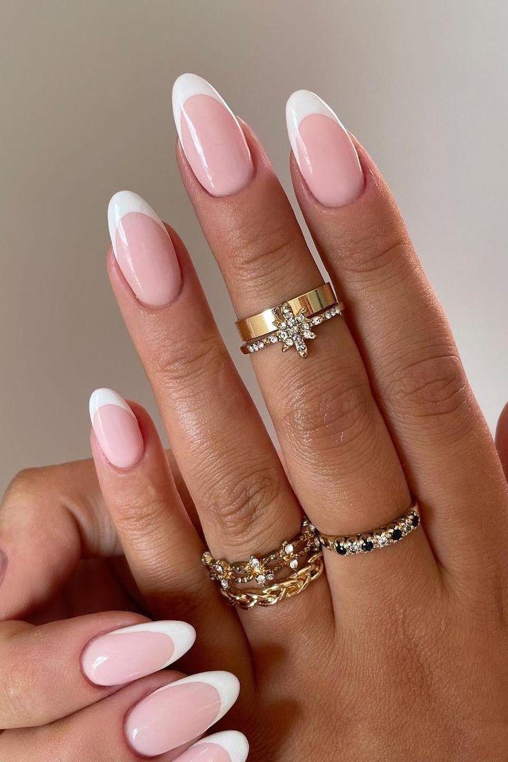 Chic Almond-Shaped French Manicure with Elegant Pink and White Design, Accentuated by Delicate Gold and Silver Rings.