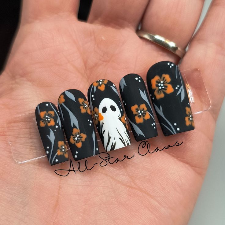 Whimsical Matte Black Nail Design with Bright Orange Floral Patterns and Playful Ghost for Fall.