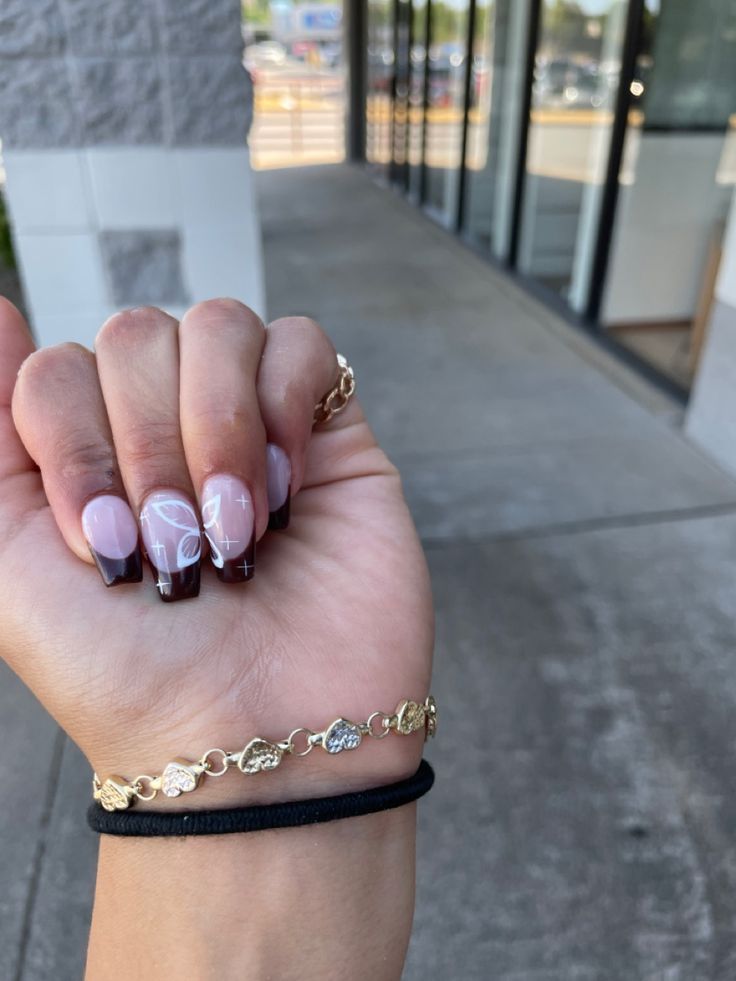 Elegant Nail Design: Soft Pink and Dark Brown Tips with White Patterns and Gold Bracelet.