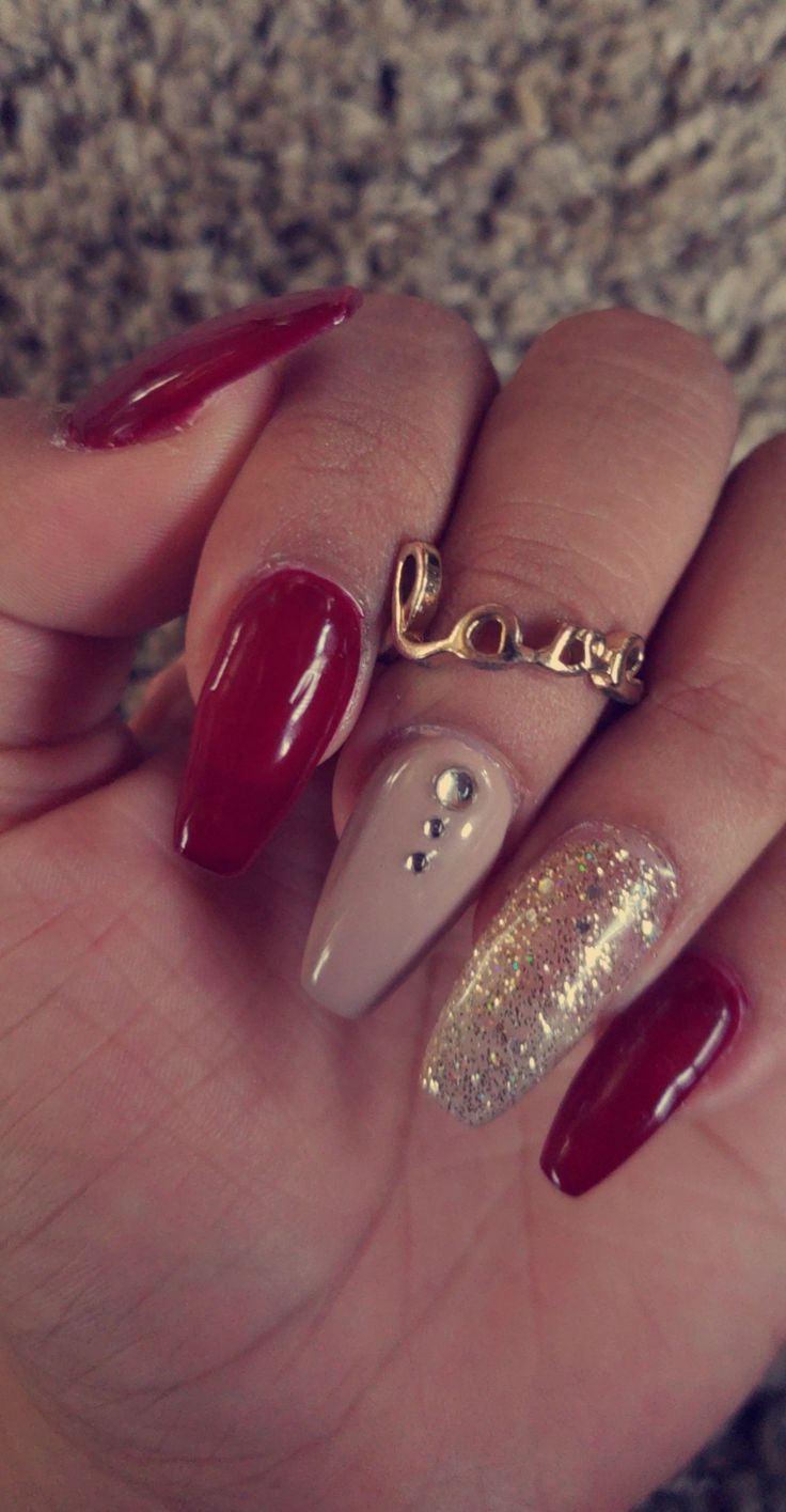 Chic Nail Design: Deep Red, Beige, and Sparkly Gold Elegance with Gems and Gold Accents.