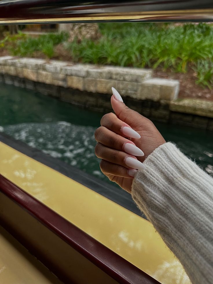 Chic Almond-Shaped Glossy White Nails Complementing a Neutral Sweater Against a Serene Waterfront.